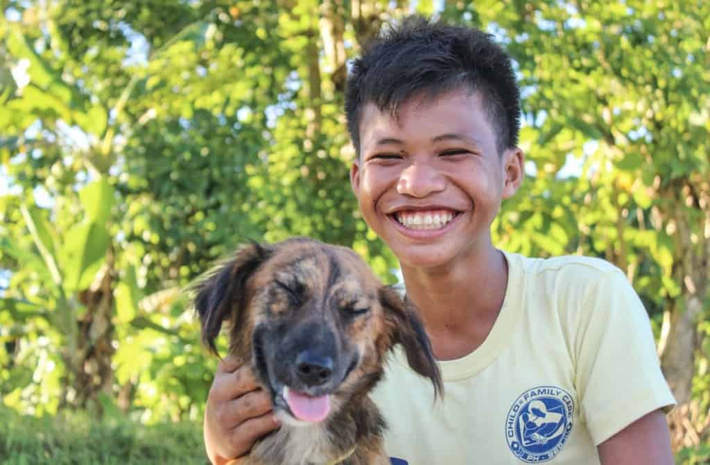 Raymark is wearing a light green shirt. He is outside with the family pet, a dog named Marga. There are trees in the background.