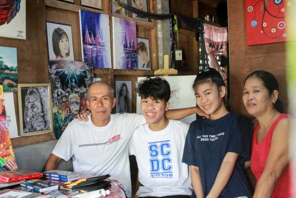 Jandel is wearing a white shirt with blue letters on it. He is sitting inside his home with his mother, father, and twin sister, Janine. Some of his paintings are hind them.