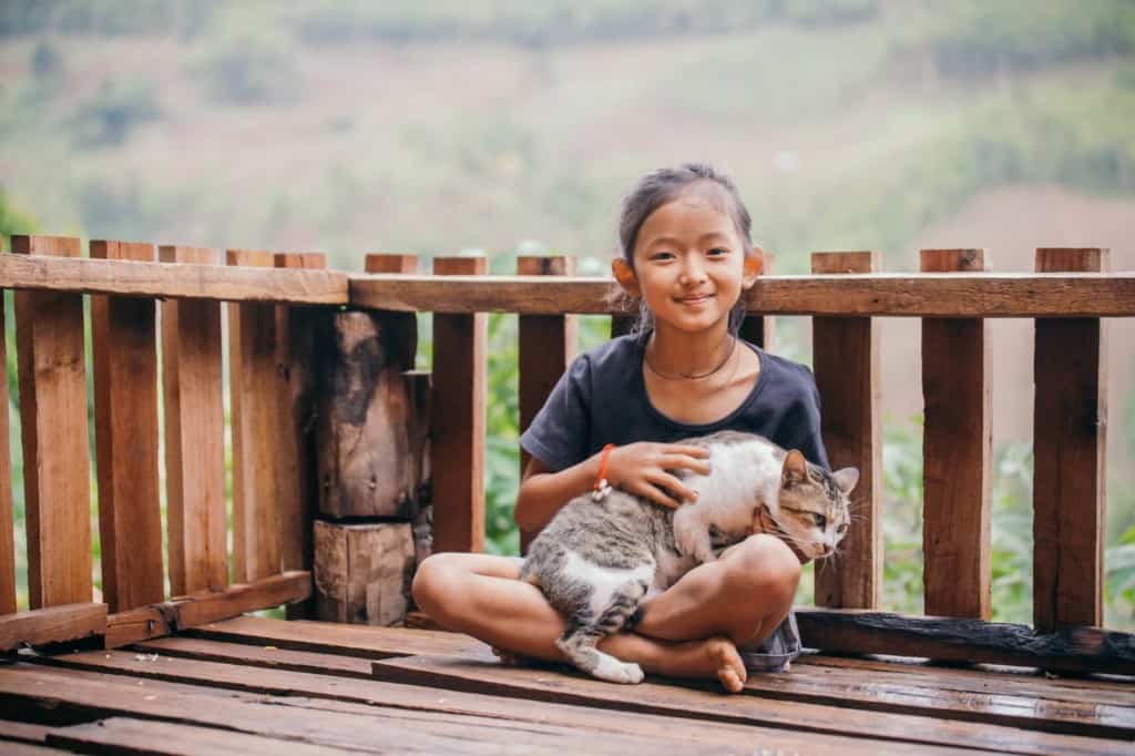 Chonticha is wearing a black shirt and shorts. She is sitting on a deck on her home and is holding her pet cat named Mama-Big.