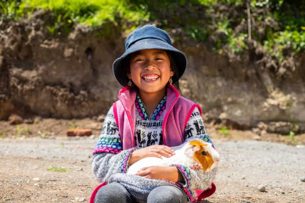 Emily is at the guinea pig farm, holding a guinea pig and smiling at the camera. She is wearing a sweater, a pink vest, and a blue hat.