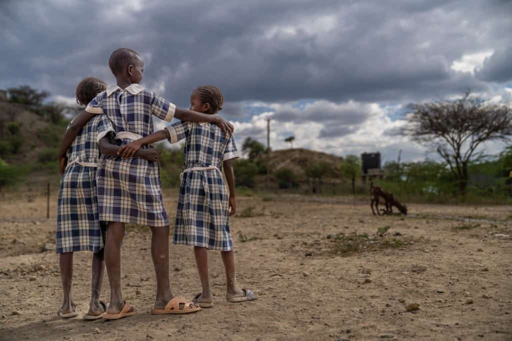 Joan is standing outsdie with two of her friends, Lodio and Chelatan. They are wearing their uniforms, blue and white plaid dresses with white collars and belts.They have their arms around each other. 