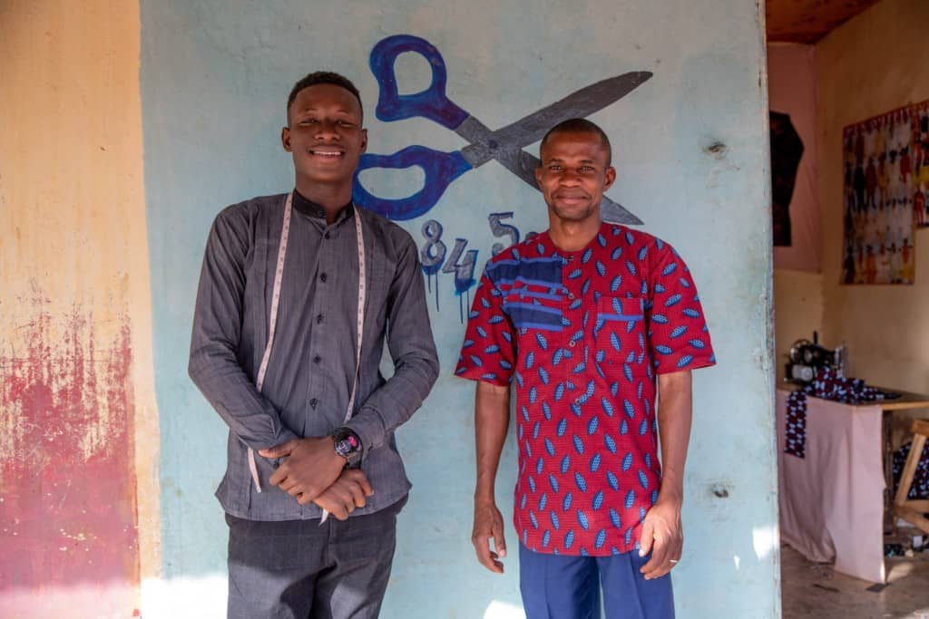 Honore is wearing a gray shirt and has a measuring tape around his neck. He is standing with his arms crossed in front of him next to Judes, the Project Director. Judes is wearing a red and blue patterned shirt. There is a pair of scissors painted on the wall behind them.