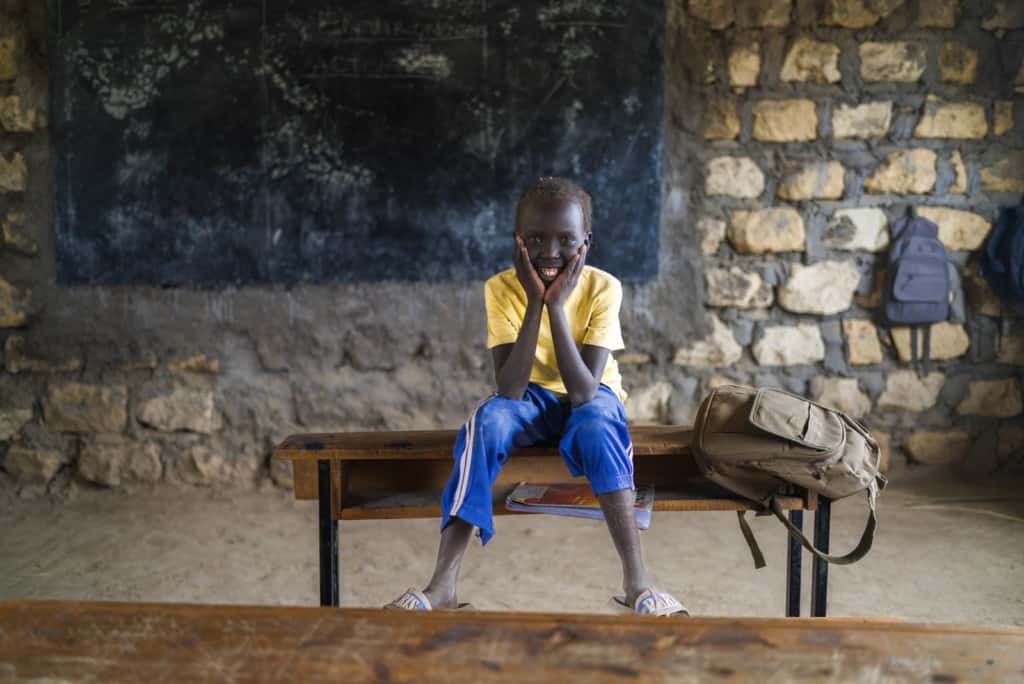 Joan is wearing a yellow shirt and blue pants. She is sitting on a bench inside the Compassion center with her chin resting in her hands.