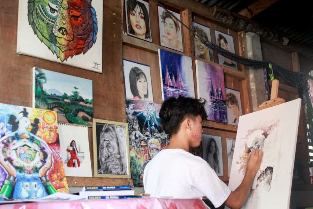 Jandel is wearing a white shirt with blue letters on it. He is sitting inside his home and is painting on a canvas that is on an easel. Some of his other paintings are propped up behind him.