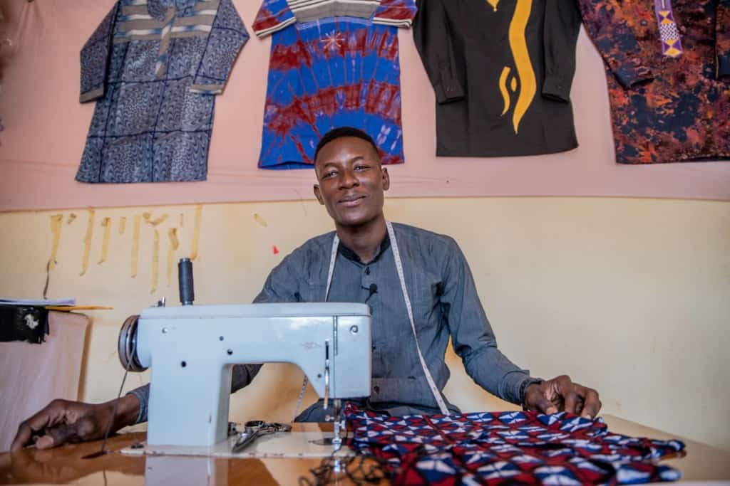 Honore is wearing a gray shirt and has a measuring tape around his neck. He is sitting at a sewing machine in his workshop and there are shirts hanging on the wall behind him.