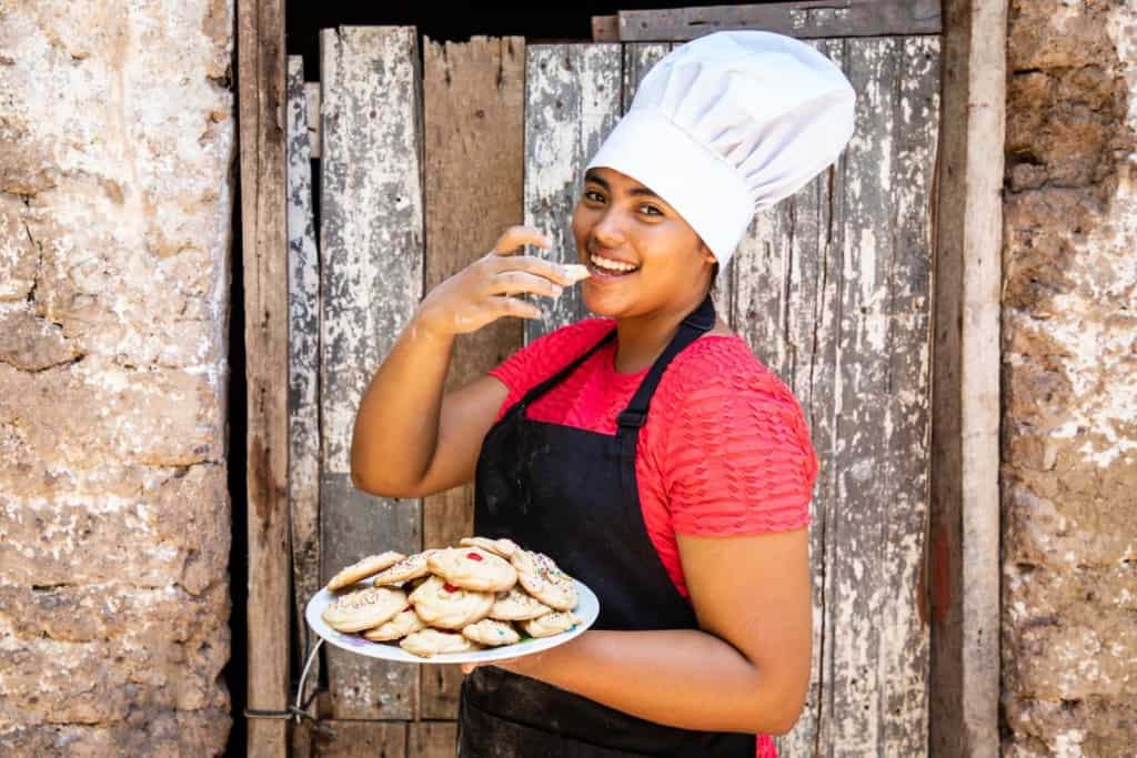 Karla is wearing a red shirt, black apron, and a chef's hat. She is standing in front of her home and is holding a plate of cookies she made. She is holding one of the cookies close to her mouth.