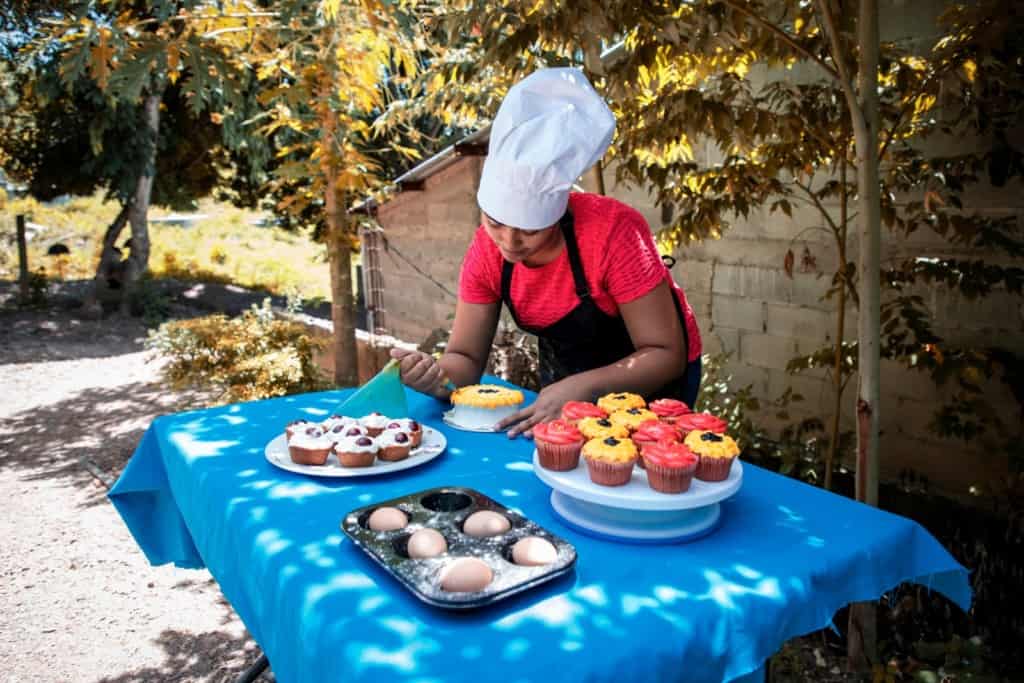 Karla is wearing a red shirt, black apron, and a chef's hat. She is decorating a cake on a table in her front yard. There are cupcakes on the table next to her.