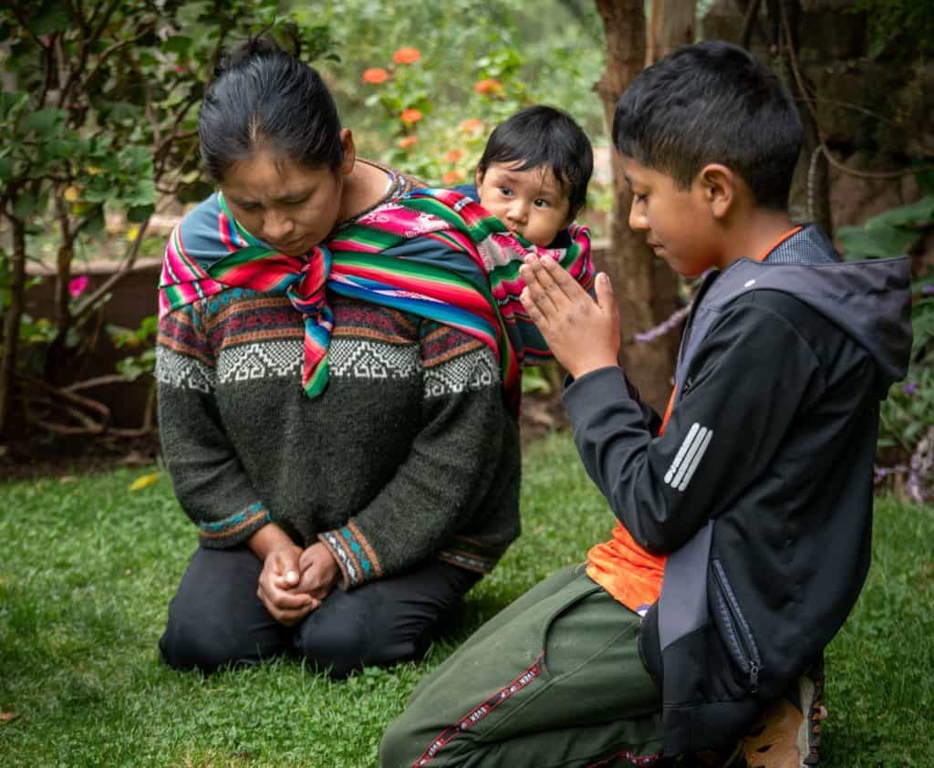 Boy wearing a black jacket with a gray hood. He is kneeling down in his yard and is praying with his mother, who is holding her youngest child on her back.