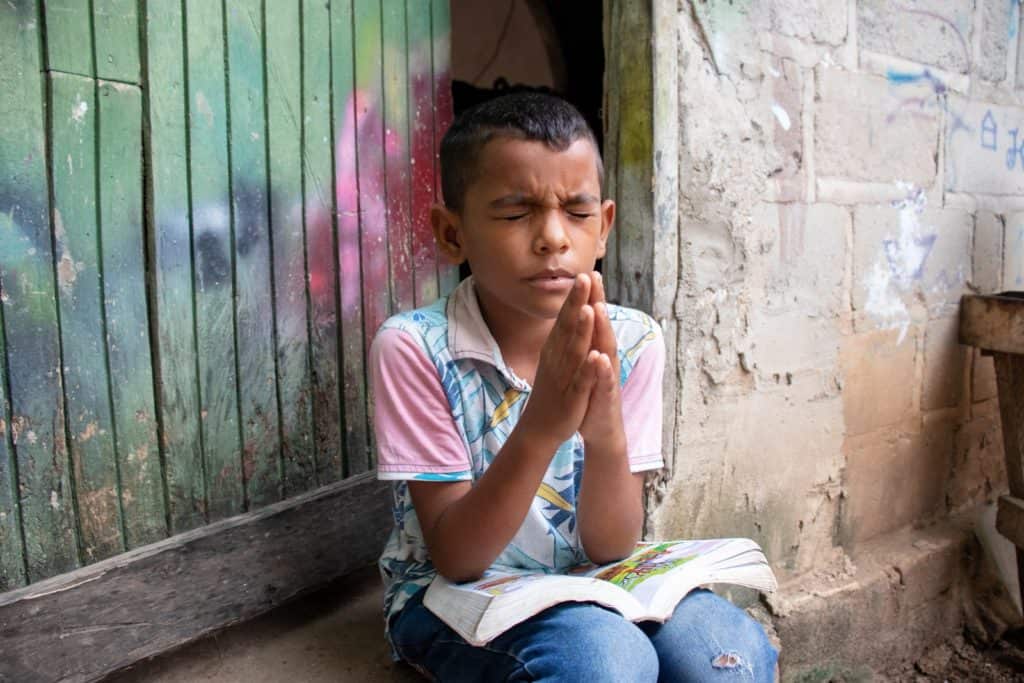 Boy wearing a blue and white patterned shirt with pink sleeves and jeans. He is sitting outside his home praying with his hands pressed together in front of him. On his lap is his Bible.