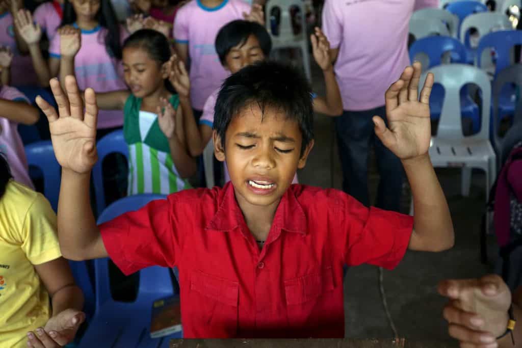 Young boy wearing a red shirt. He is praying with a group of children in the background. He has both hands in the air.
