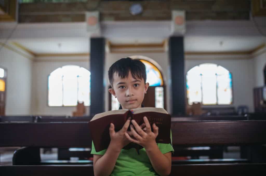 Boy wearing a bright green dress with a black stripe across the front. He is sitting in a church pew and is holding a Bible.