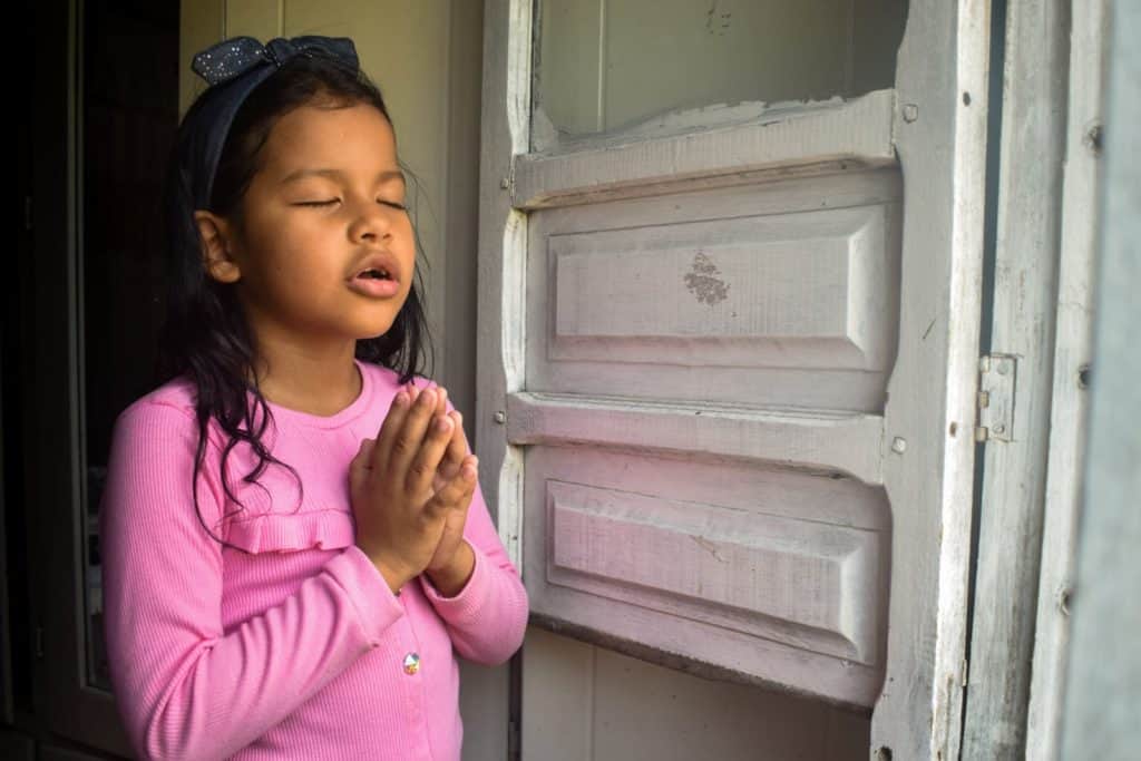 Girl wearing a pink shirt. She is inside her home and is praying. She has her hands folded in front of her chest.