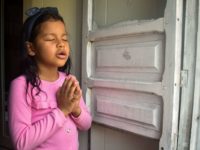 Girl wearing a pink shirt. She is inside her home and is praying. She has her hands folded in front of her chest.