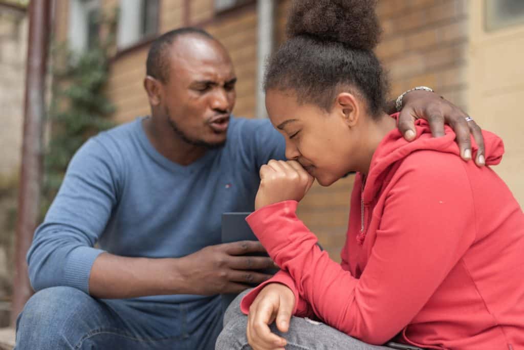 Man wearing a blue shirt and is praying with a girl wearing a red sweatshirt. His hand is on her back.