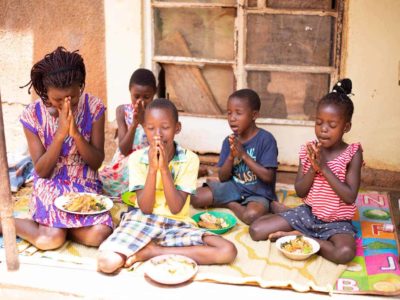 Raphael is wearing a yellow shirt with blue, white, and yellow plaid shorts. He is siting outside his home with his mother and siblings. They are praying before they eat. Each of them has a bowl of food in front of them.