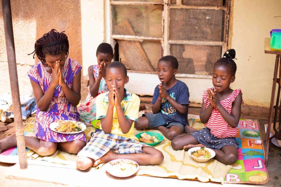 Raphael is wearing a yellow shirt with blue, white, and yellow plaid shorts. He is siting outside his home with his mother and siblings. They are praying before they eat. Each of them has a bowl of food in front of them.