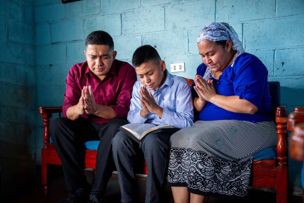 Josué is wearing a blue and white striped shirt. He is sitting inside his home on a couch with his mother, wearing a blue shirt, and his father, wearing a maroon shirt. They are all praying together. The wall behind them is blue.