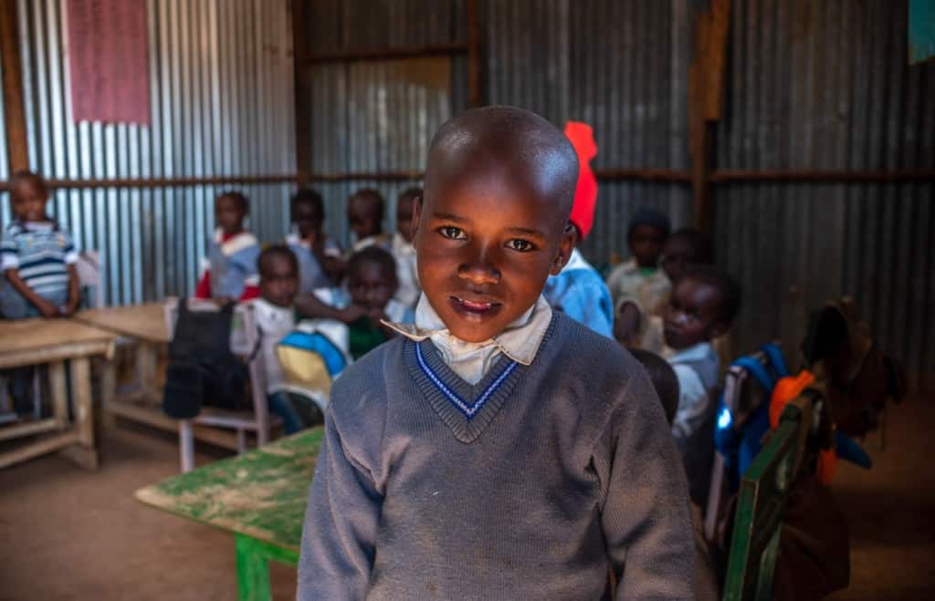 John is looking at the camera, smiling. He is wearing a gray sweater. There are several children in the background.