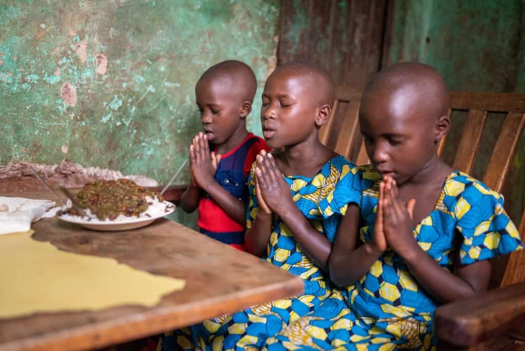 Girl wearing a red and blue dress. She is sitting at a table inside her home with her foster sister. They are praying before they eat.