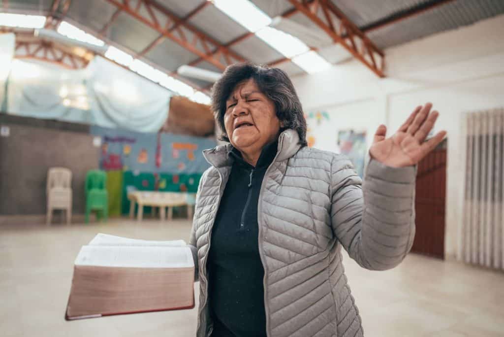 Rosmary, in a gray jacket, is holding a Bible in one hand and is raising the other one. She is praying, and her eyes are closed.