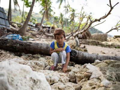 A boy wearing a blue and yellow shirt sits among wood and rocks in the Philippines.