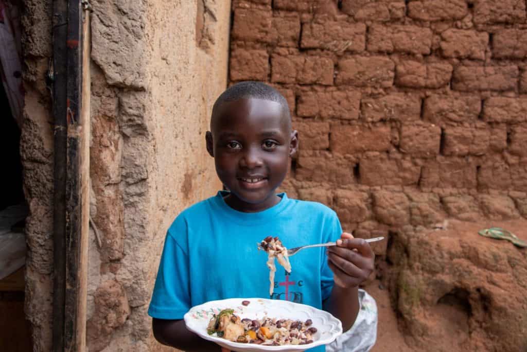 Girl, wearing a blue shirt, is holding a plate of food her mother prepared for her. She is standing outside her home.