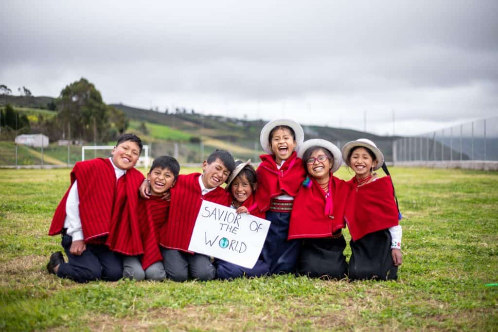 All seven children are wearing traditional clothing. They are kneeling down outside and one of the children is holding a signi that says, "Savior of the World."