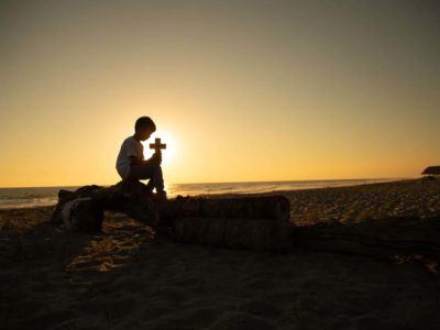 Boy sitting on a rock at the beach and is holding a cross.