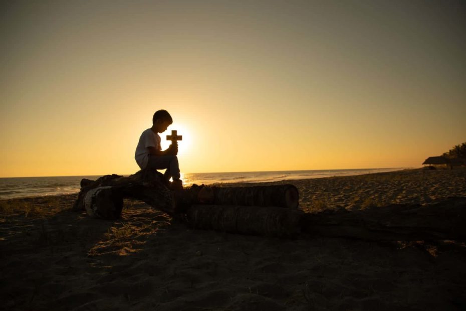 Boy sitting on a rock at the beach and is holding a cross.