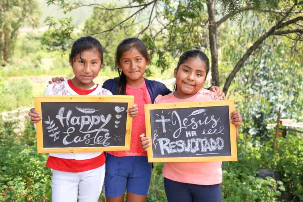 Three girls stand together outside. Two of the girls are holding hand written chalk board signs, "Happy Easter and Jesus Resucitado, Jesus is Risen."