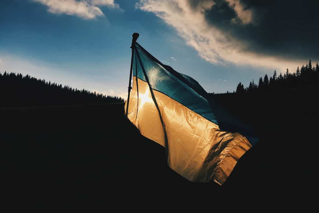 The blue and yellow Ukrainian flag is flying against a backdrop of trees, blue sky and clouds. The sun is shining behind it.