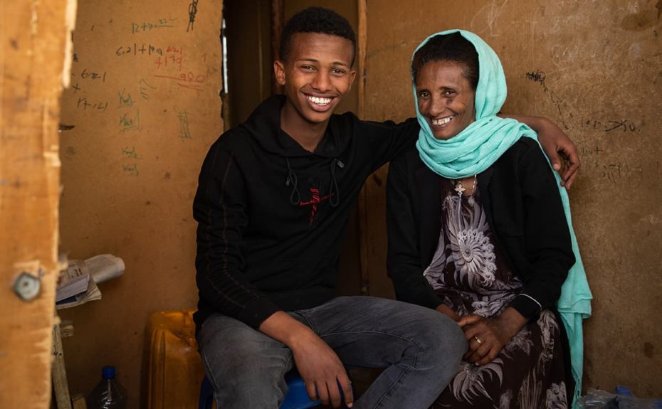 Mikiyas, wearing a black shirt and blue jeans, is sitting next to his mother with his arm around her. She is wearing a teal blue head wrap and a black and white dress. They are smiling.