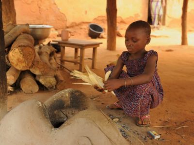 Girl squatting by a fire holding a corn husk.