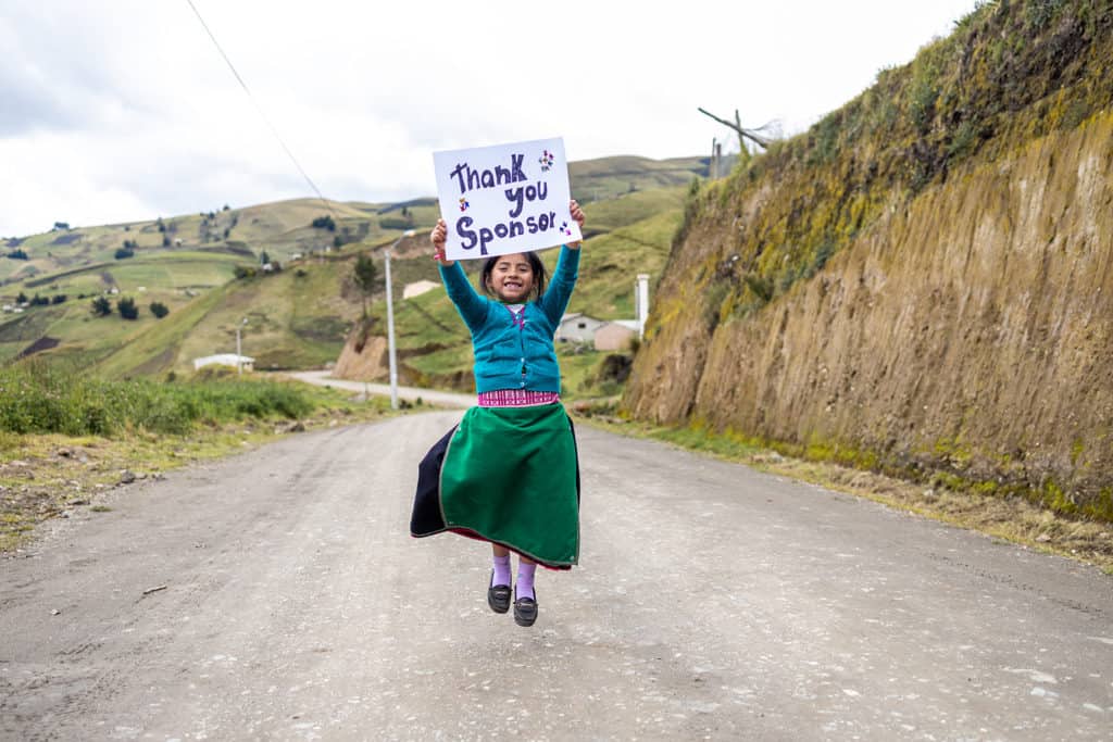 Lizeth holds a sign saying "Thank you sponsor" as she jumps into the air on a rural road in Ecuador. There are grassy hills in the background. Lizeth is wearing a green skirt and blue sweater.
