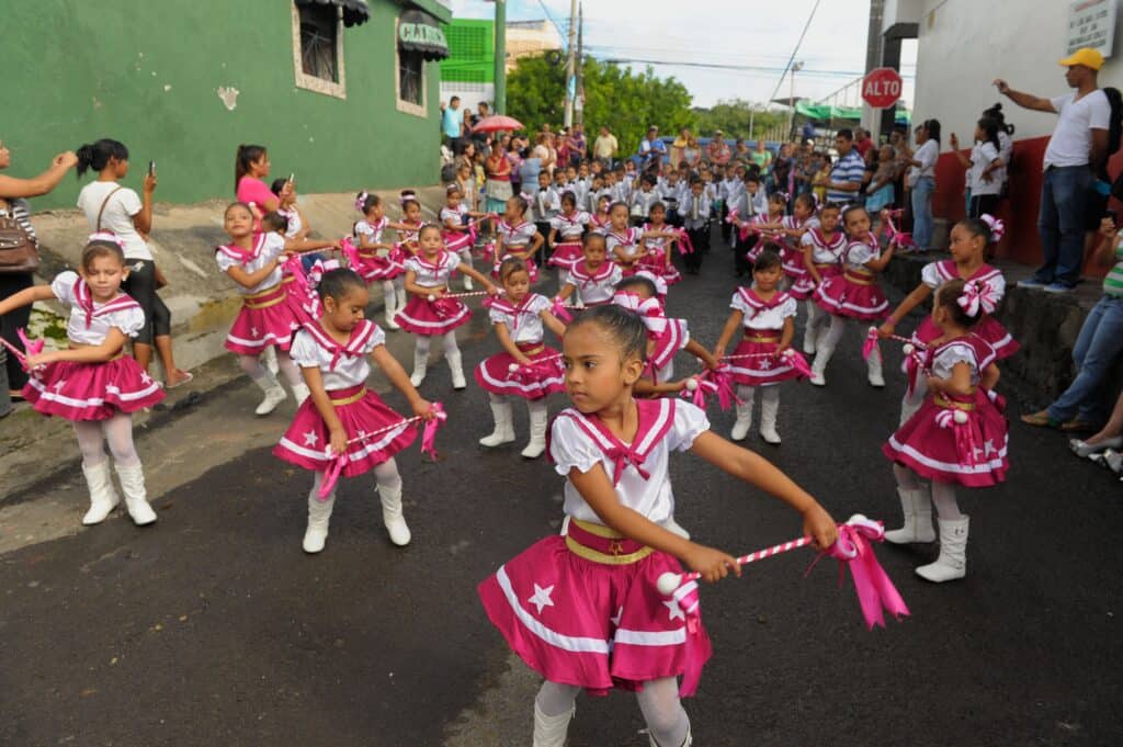 Children clothed in bright white and pink outfits twirl batons down a street in El Salvador