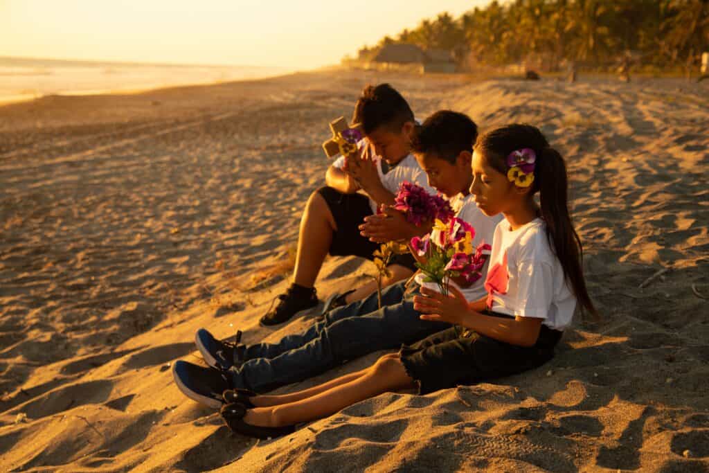 Children in El Salvador sit on the beach holding flowers and a decorated cross as they pray