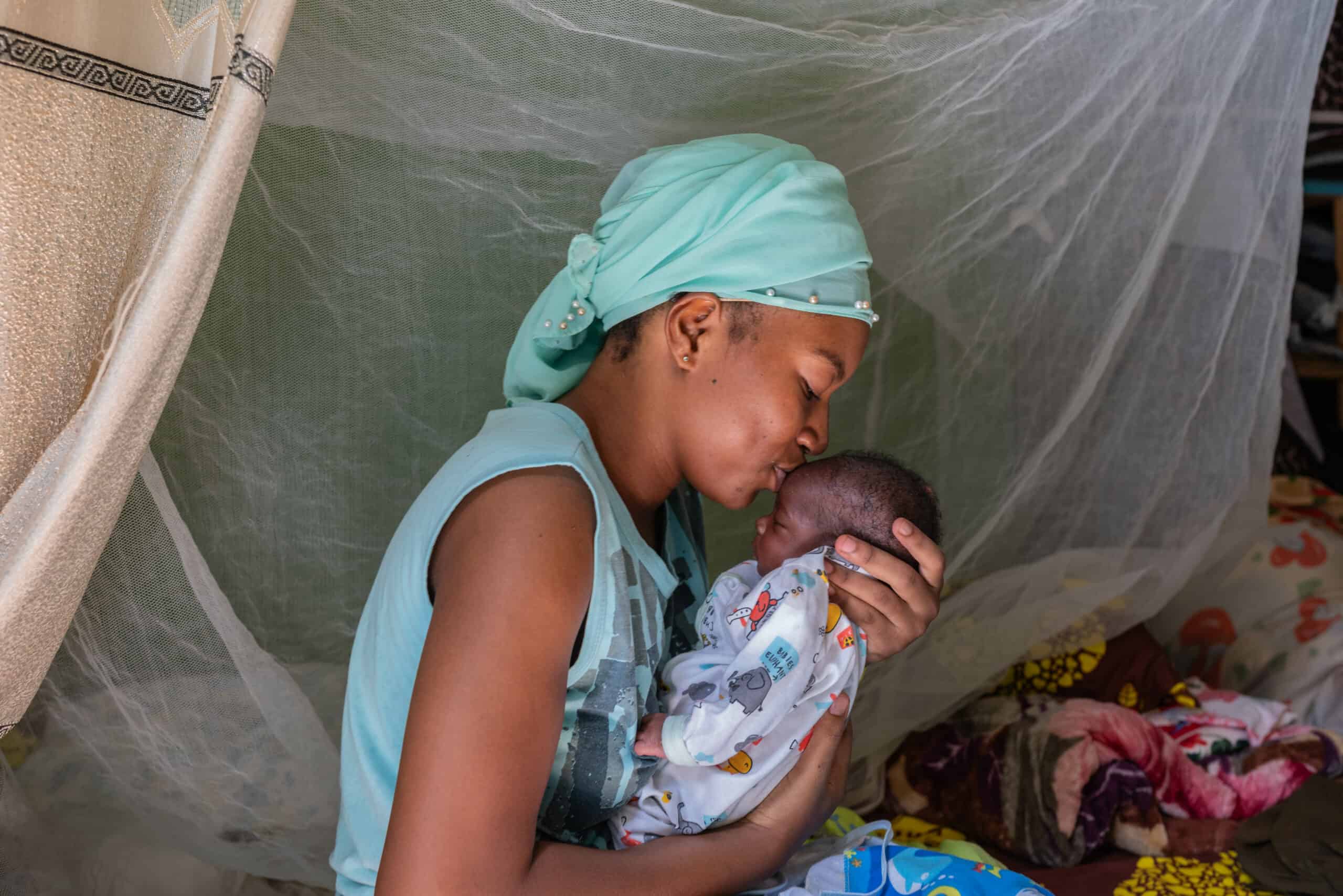 A mom wearing a light blue head wrap kisses her infant child on the forehead.