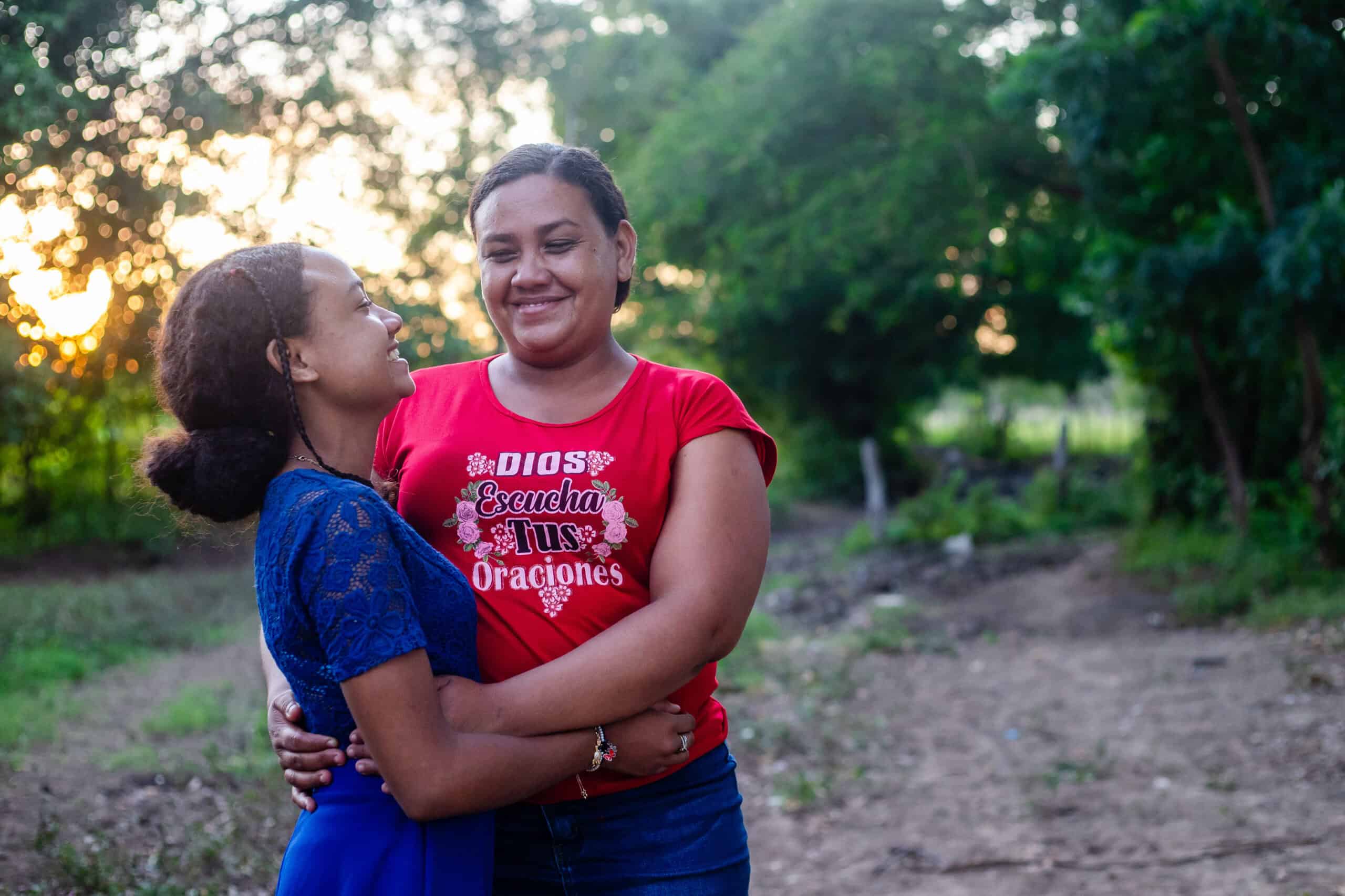 A young girl wearing a blue dress hugs her mother as they smile at each other.