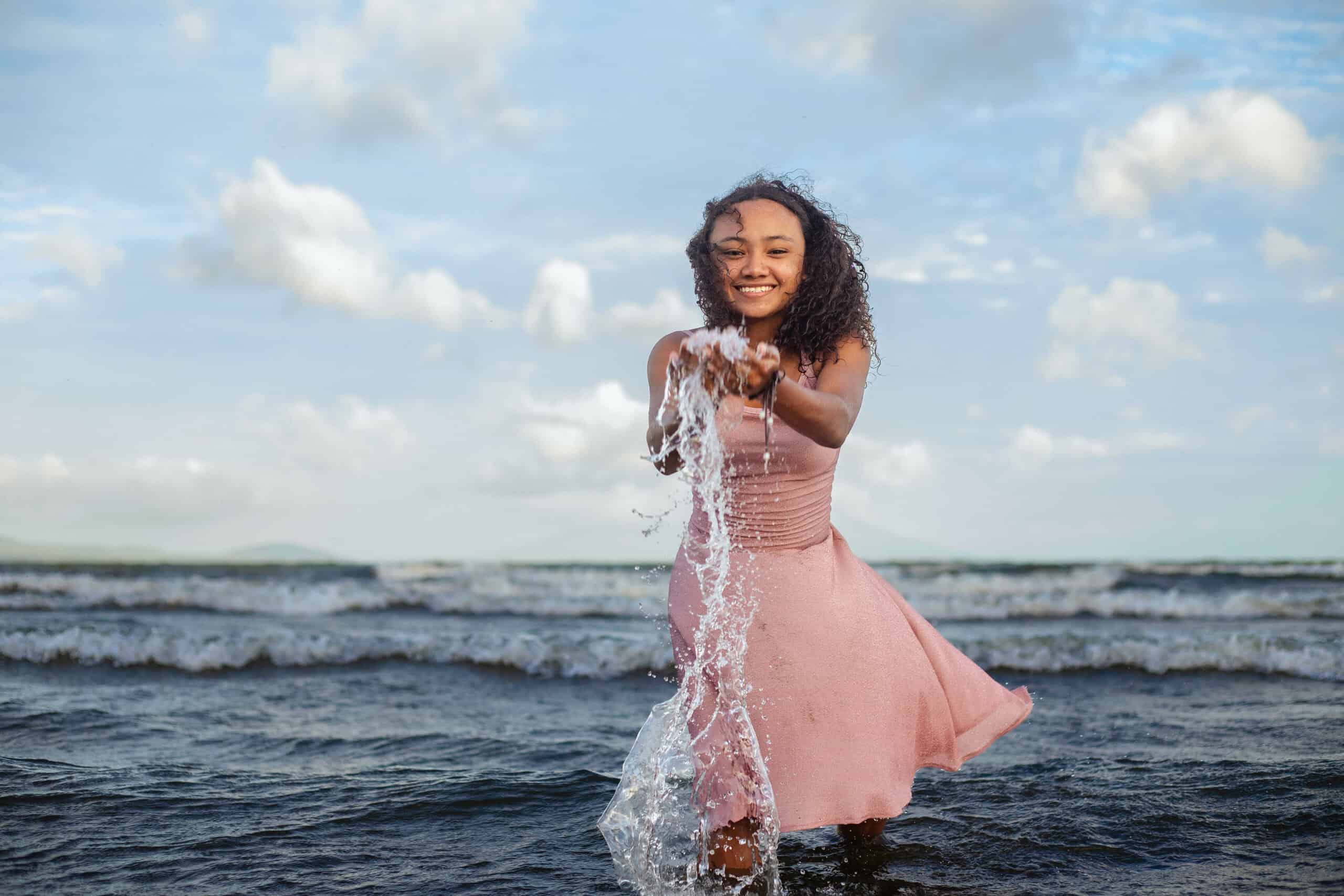 A young girl wearing a pink dress stands in a lake with water flowing out of her hands as she smiles for the camera.