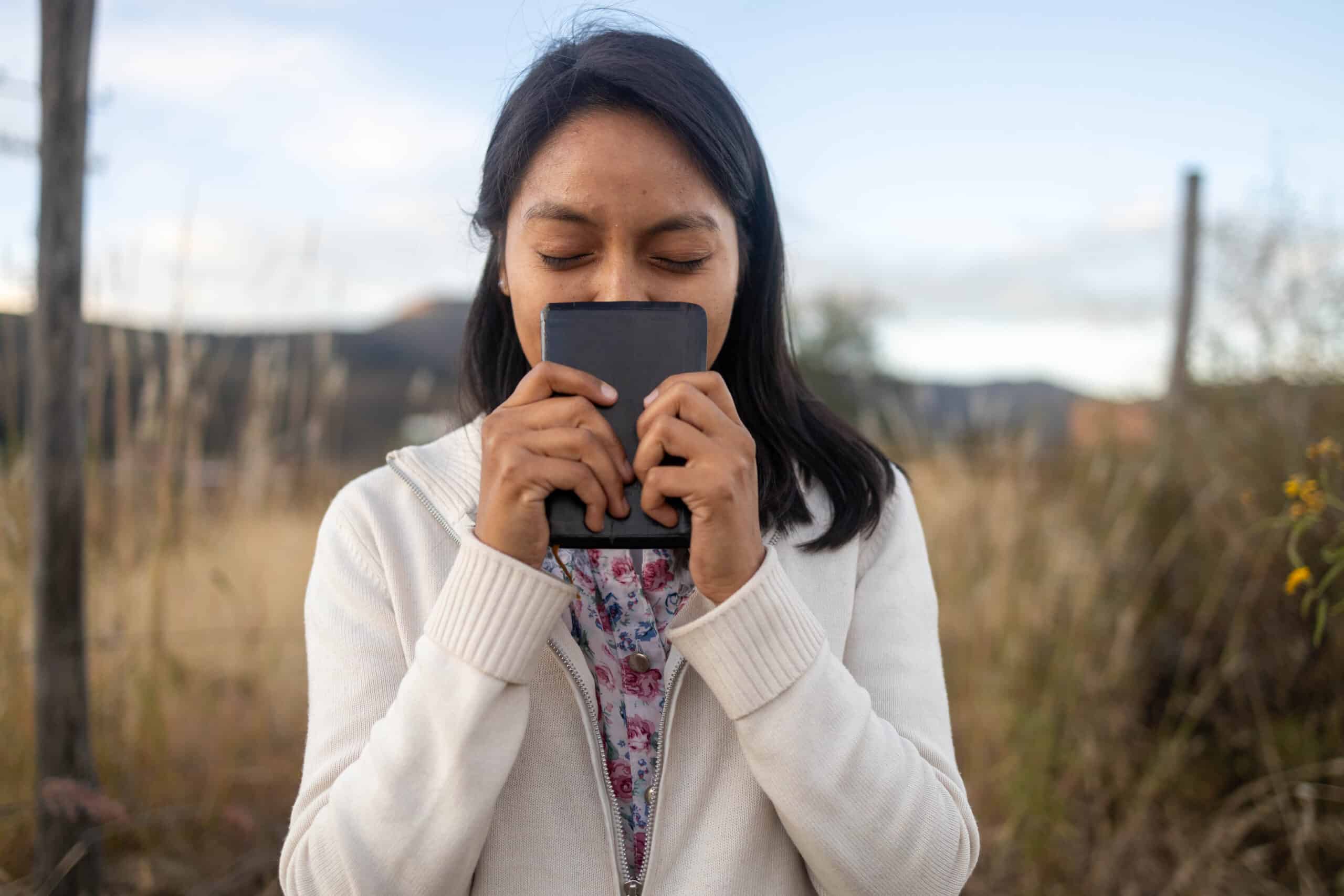 A girl wearing a cream sweater holds a bible to her face with her eyes closed.