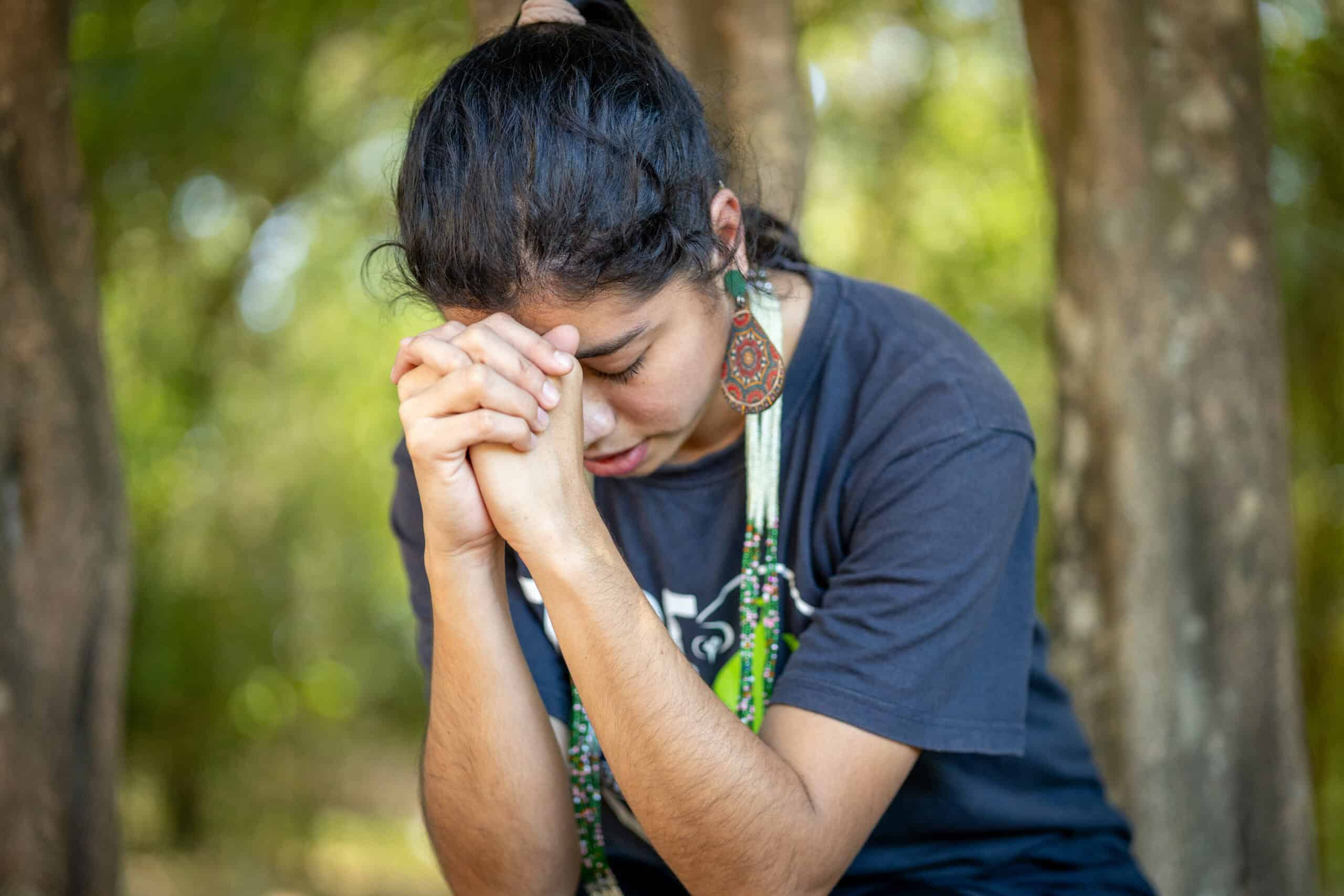 A young woman wearing beaded earrings and a necklace prays with her hands folded and head bowed.