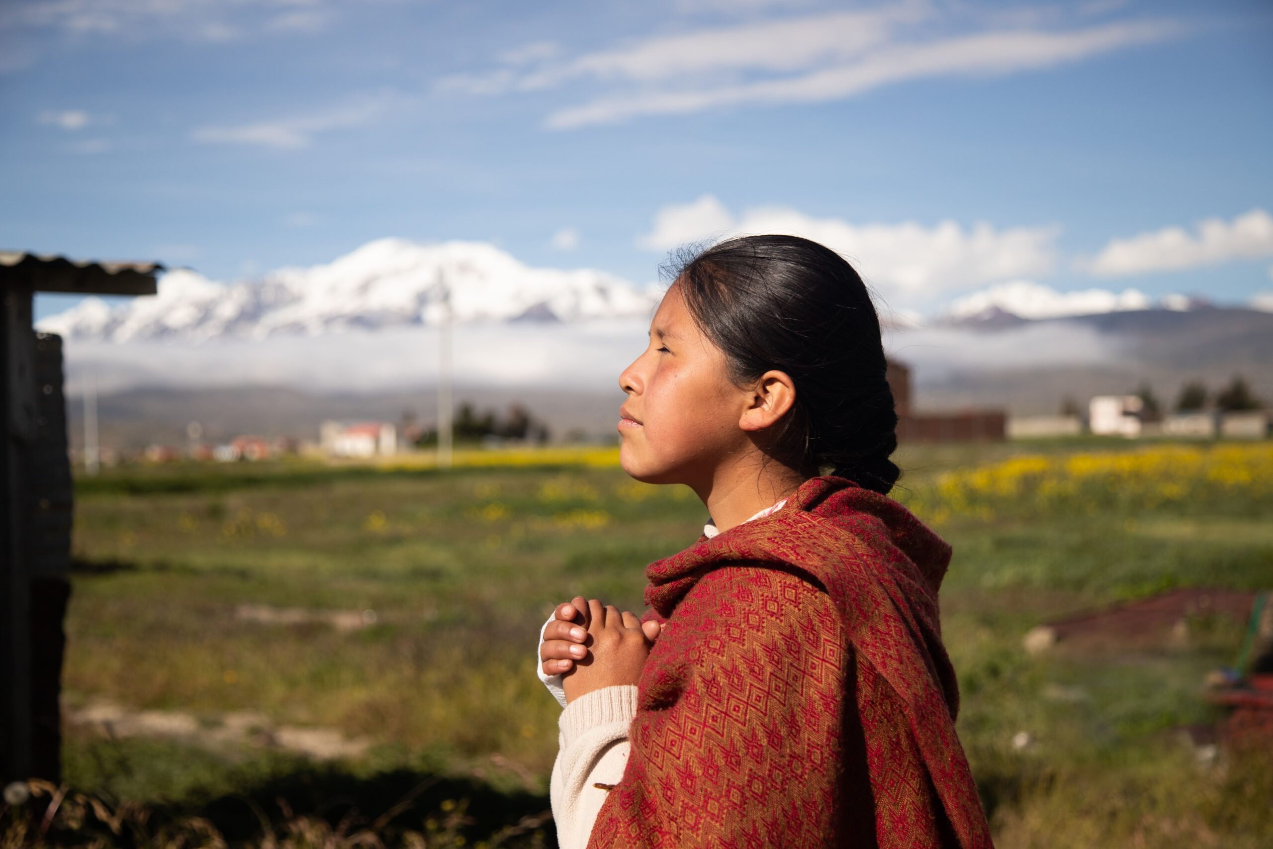 A young woman wears a shawl and stands with her eyes closed and hands clasped together as she prays.