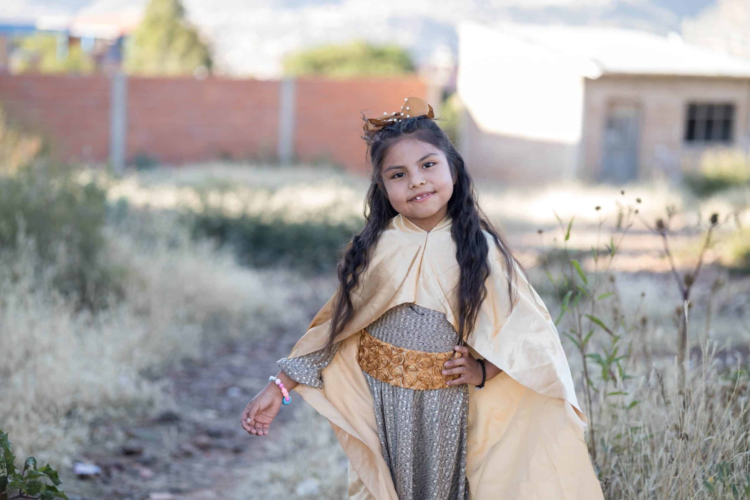 A young girl wears a gold bow, robe and belt while smiling for the camera in a field of grass.