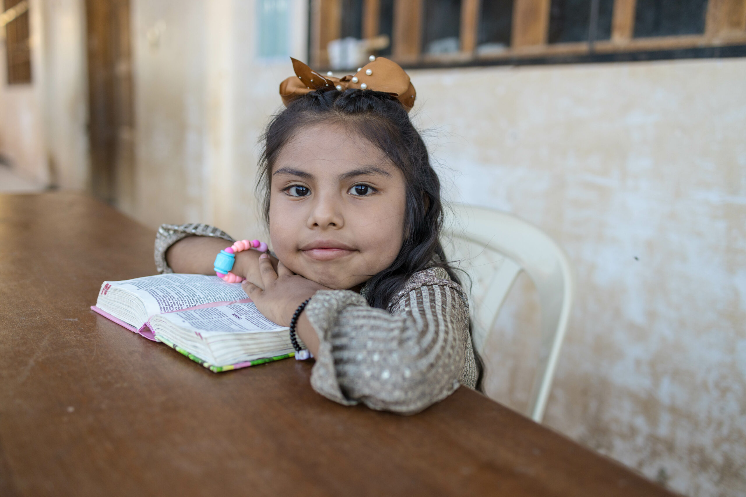 A young girl sits at a wooden table wearing a gold bow with a Bible in front of her. Her arms rest on top of the Bible and she smiles for the camera.