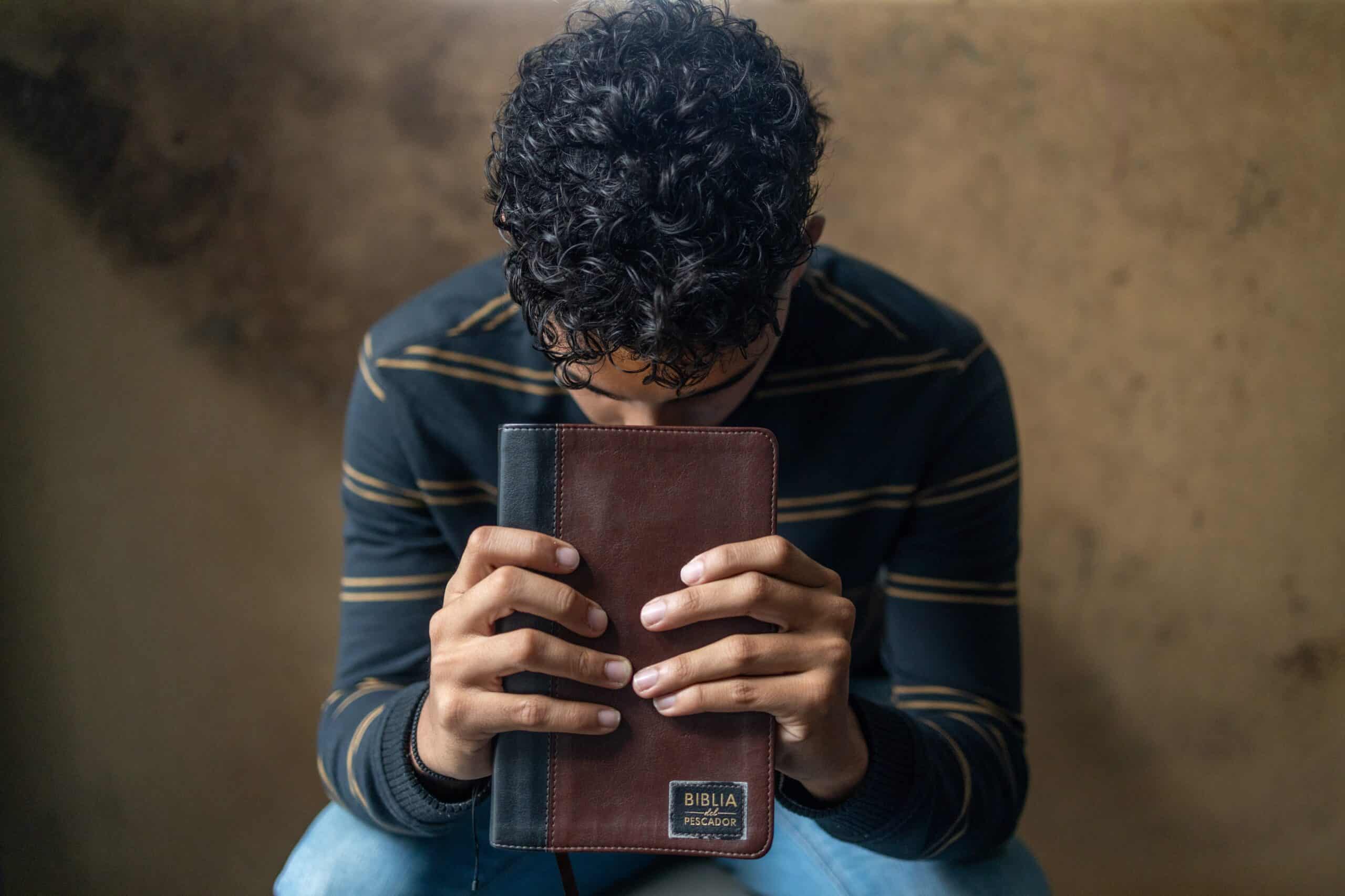 Young man wearing a striped shirt holds a Bible with his head bowed.