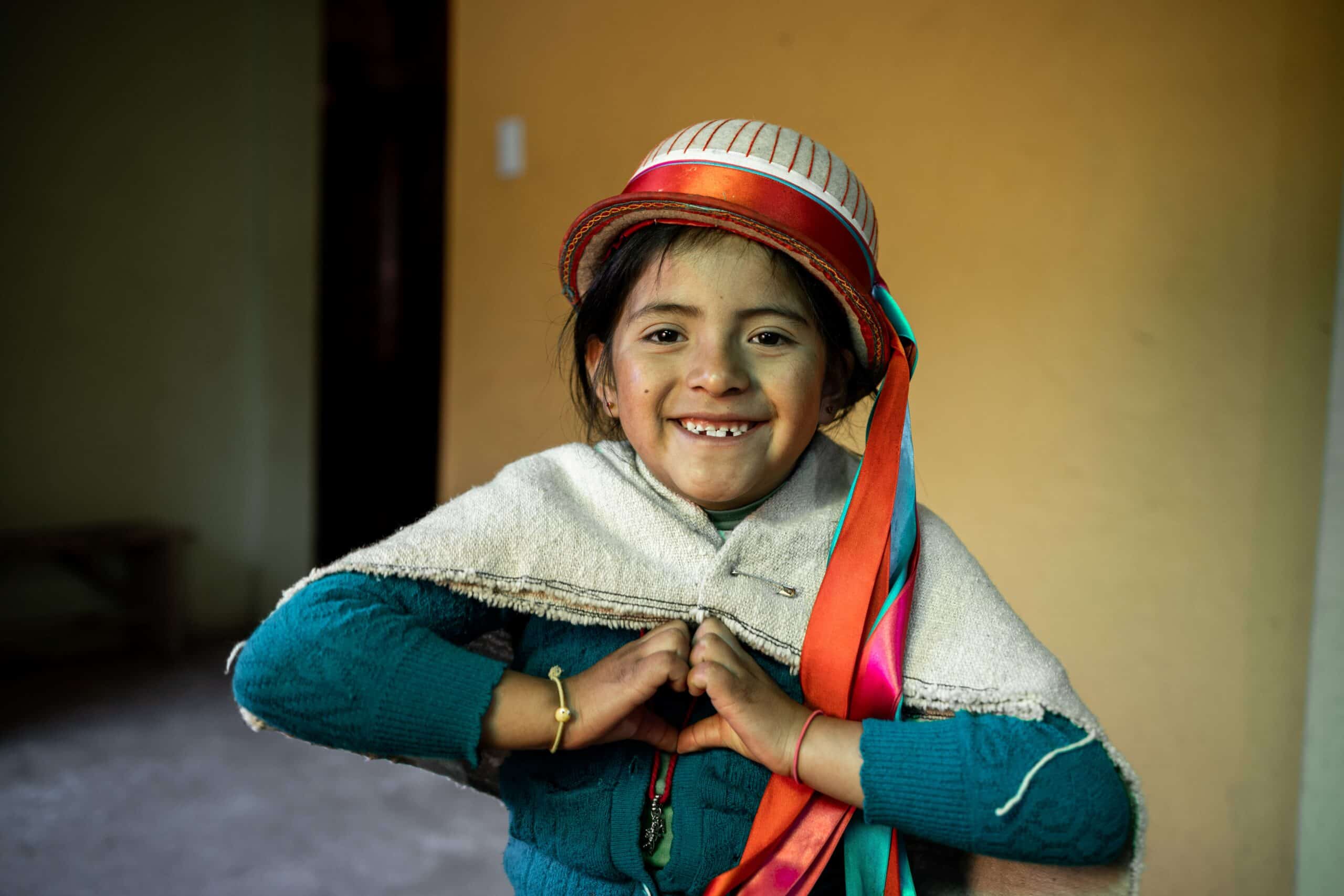 Young girl wearing a hat holds her hands together in the shape of a heart across her chest.