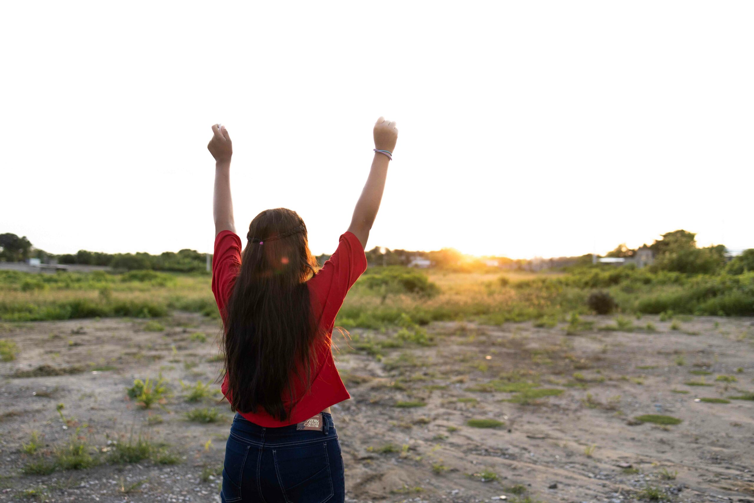 A girl with long hair wearing a red shirt raises her arms to the sky with the sunset in the background.