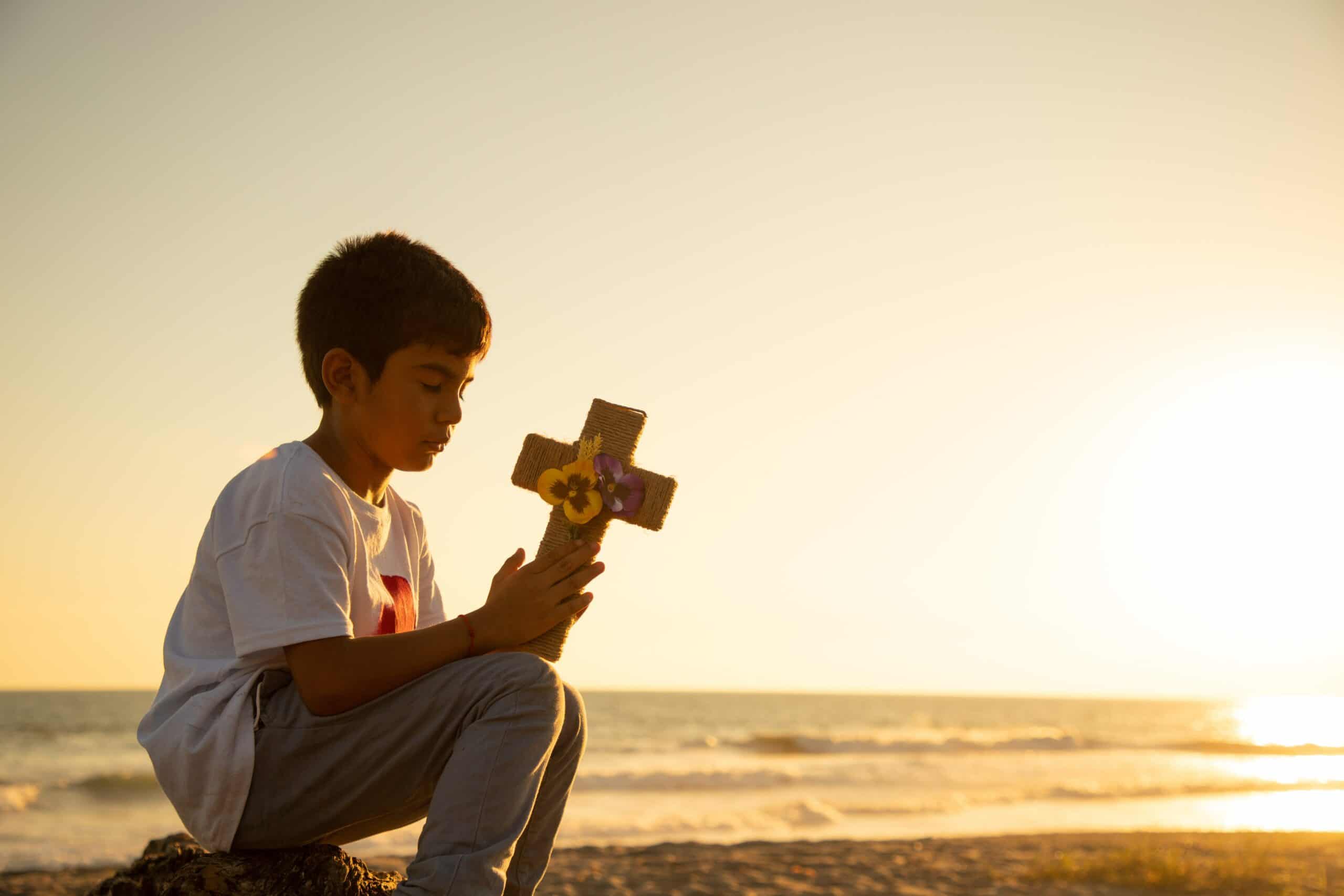 A young boy sits on a beach with the sunset in the background while holding a wooden cross and praying.