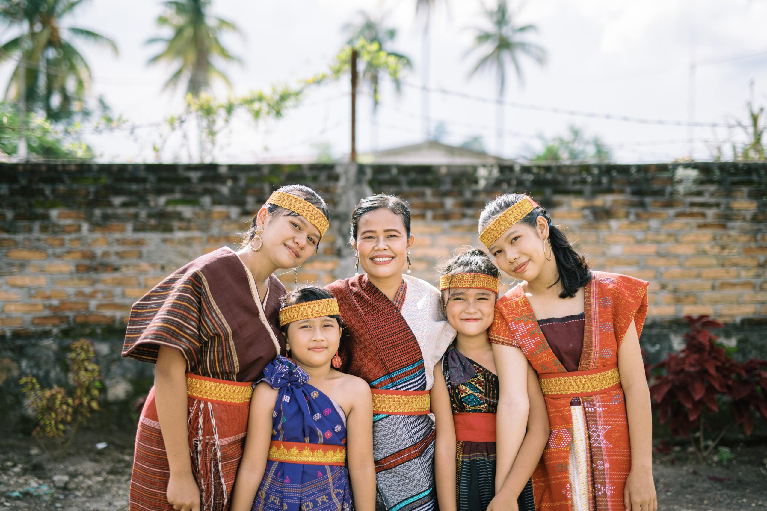 A group of women and girls wear brightly colored robes and headbands while smiling for the camera in front of a brick wall.