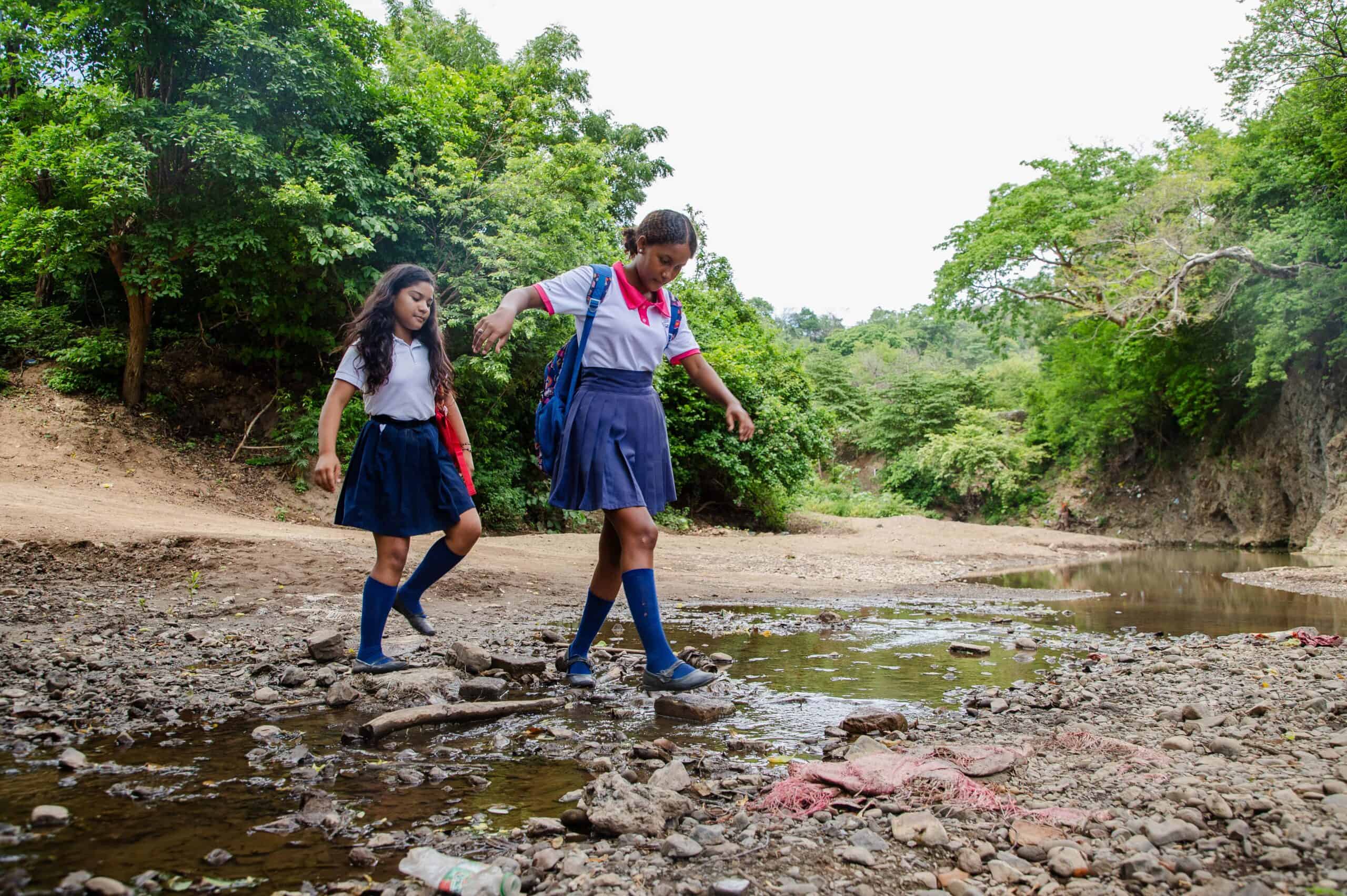 Two young girls wearing school uniforms cross a creek while smiling.